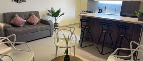 Un salon moderne avec un canapé gris, des chaises blanches, une table basse et un coin cuisine doté d’un bar et de tabourets.