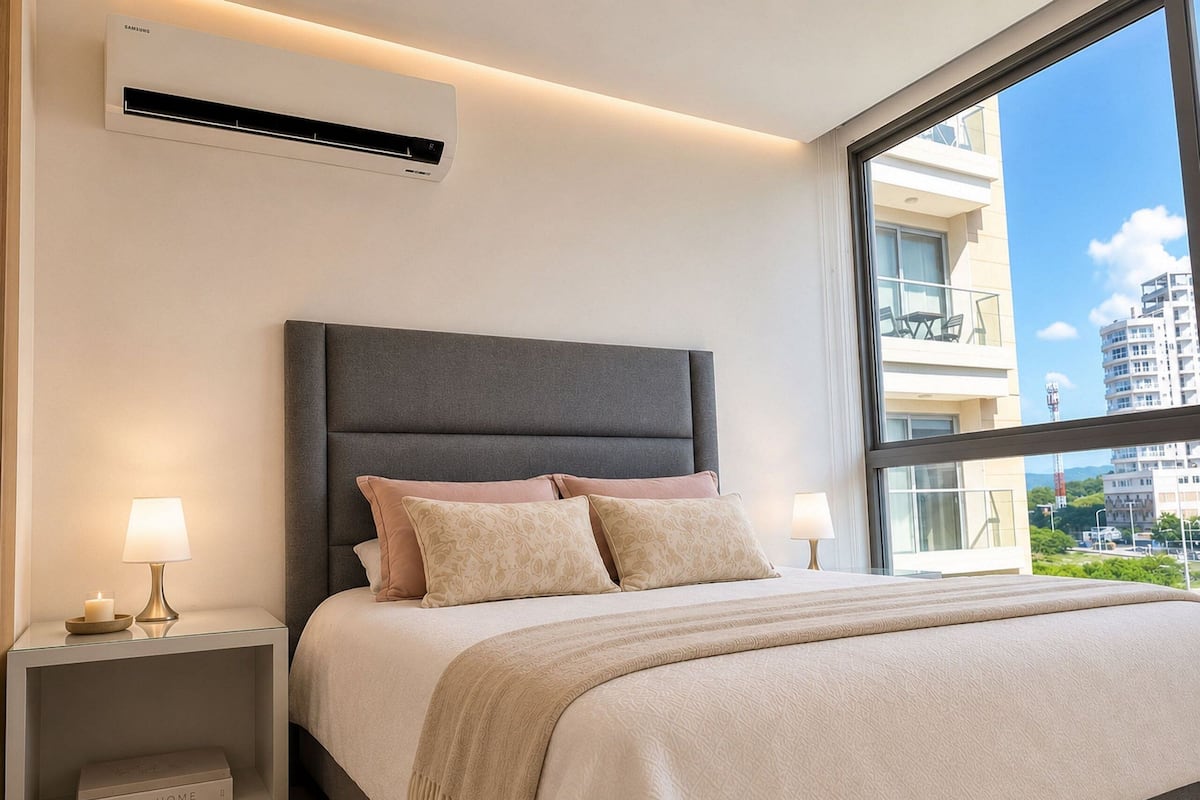 A modern bedroom with a large bed, bedside lamps, a wall-mounted air conditioner, and a view of the city through a large window.