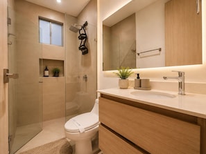 A modern bathroom with a glass shower enclosure, a toilet, a sink with a mirror above, and a small window. - Apartamentos Salguero Suites - by SOHO (Santa Marta)