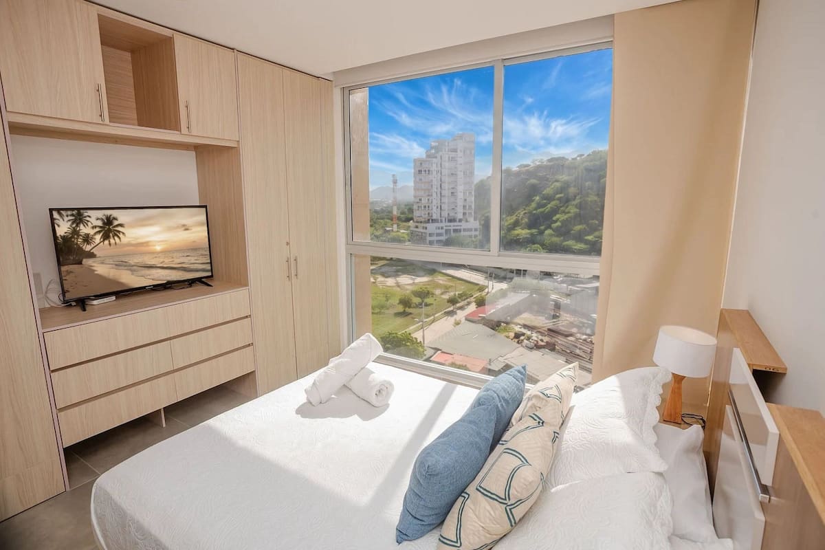 A hotel room with a bed, a TV on a stand, a window with a city view, and a wooden wardrobe.