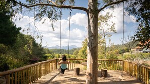 A wooden bridge with a rope swing, surrounded by trees and a mountainous landscape.