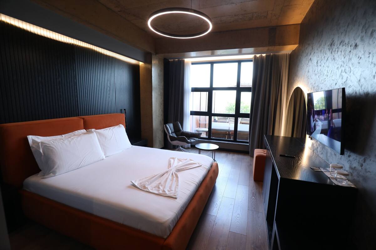 Une chambre d’hôtel avec un grand lit, une petite table, une chaise, une télévision et une fenêtre avec des rideaux.