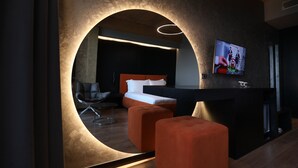 A modern hotel room with a circular wall feature, a bed with orange bedding, a black sofa, and a TV mounted on the wall. - Toka Hotel Restaurant (Udënisht)