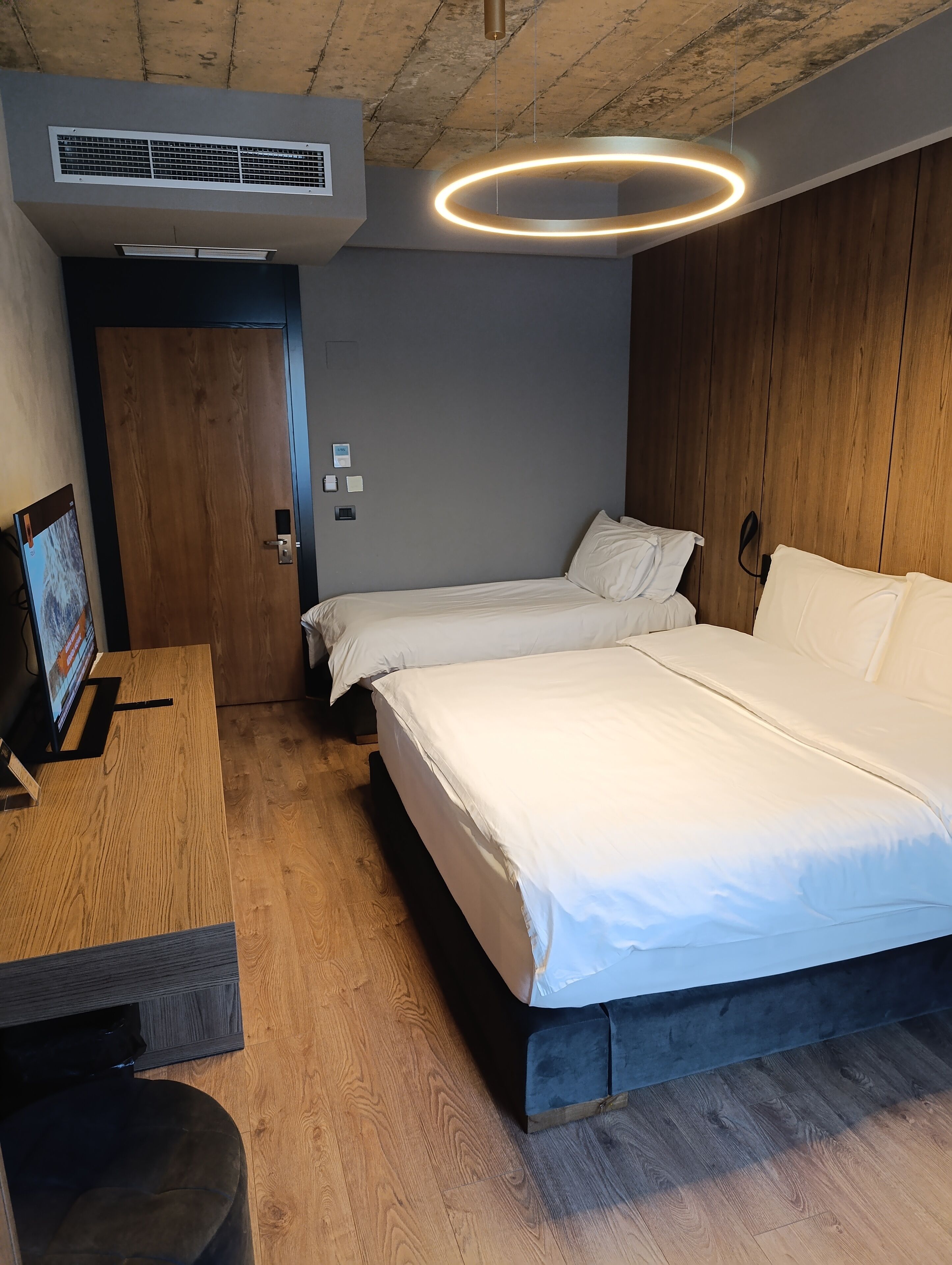 A modern hotel room with two beds, a wooden desk, a TV, and a ceiling light fixture.