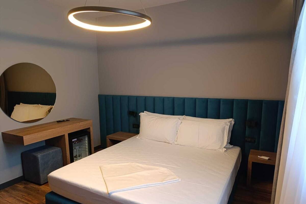 Une chambre d’hôtel moderne avec un grand lit, un miroir rond, un bureau en bois et une tête de lit bleue recouverte de tissu.