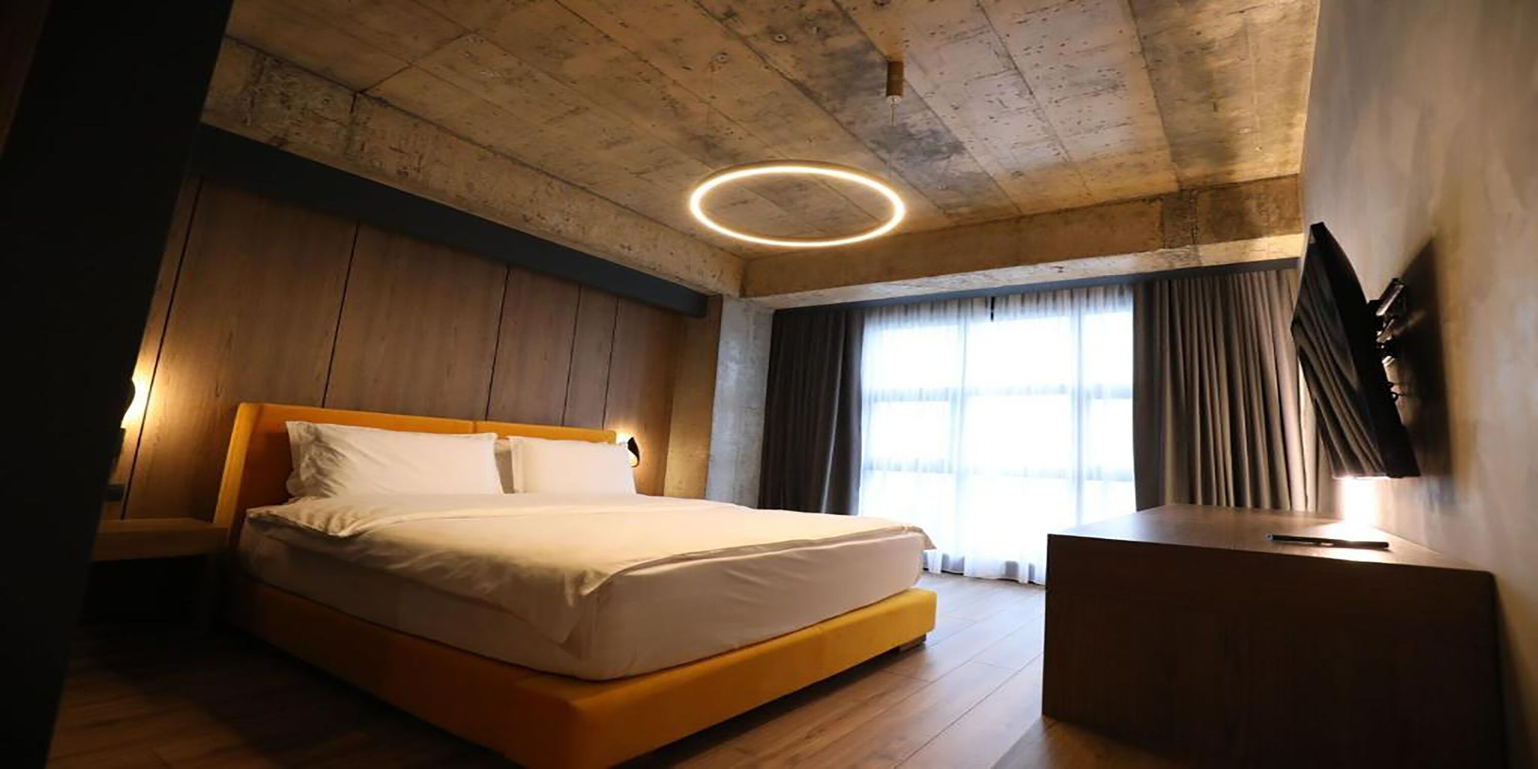 Une chambre d’hôtel moderne dotée d’un grand lit, de murs recouverts de panneaux en bois, d’un luminaire circulaire, d’un téléviseur à écran plat et d’une grande fenêtre avec des rideaux.