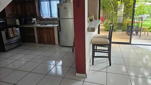 A kitchen with a refrigerator, stove, and cabinets. There is a window with blue curtains and a view of a garden outside. - APARTA HOTEL PALMEROLA (Comayagua)