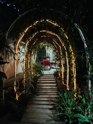 A wooden pathway leading through an archway adorned with string lights, surrounded by lush greenery. - APARTA HOTEL PALMEROLA (Comayagua)