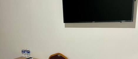 Ein Zimmer mit einem an der Wand befestigten Flachbildfernseher, einem Klapptisch mit Stuhl und einer Wasserflasche auf dem Tisch.
