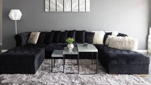 Una sala de estar moderna con un sofá seccional negro, dos otomanas negras, una mesa de centro de vidrio y una alfombra de pelo alto gris.