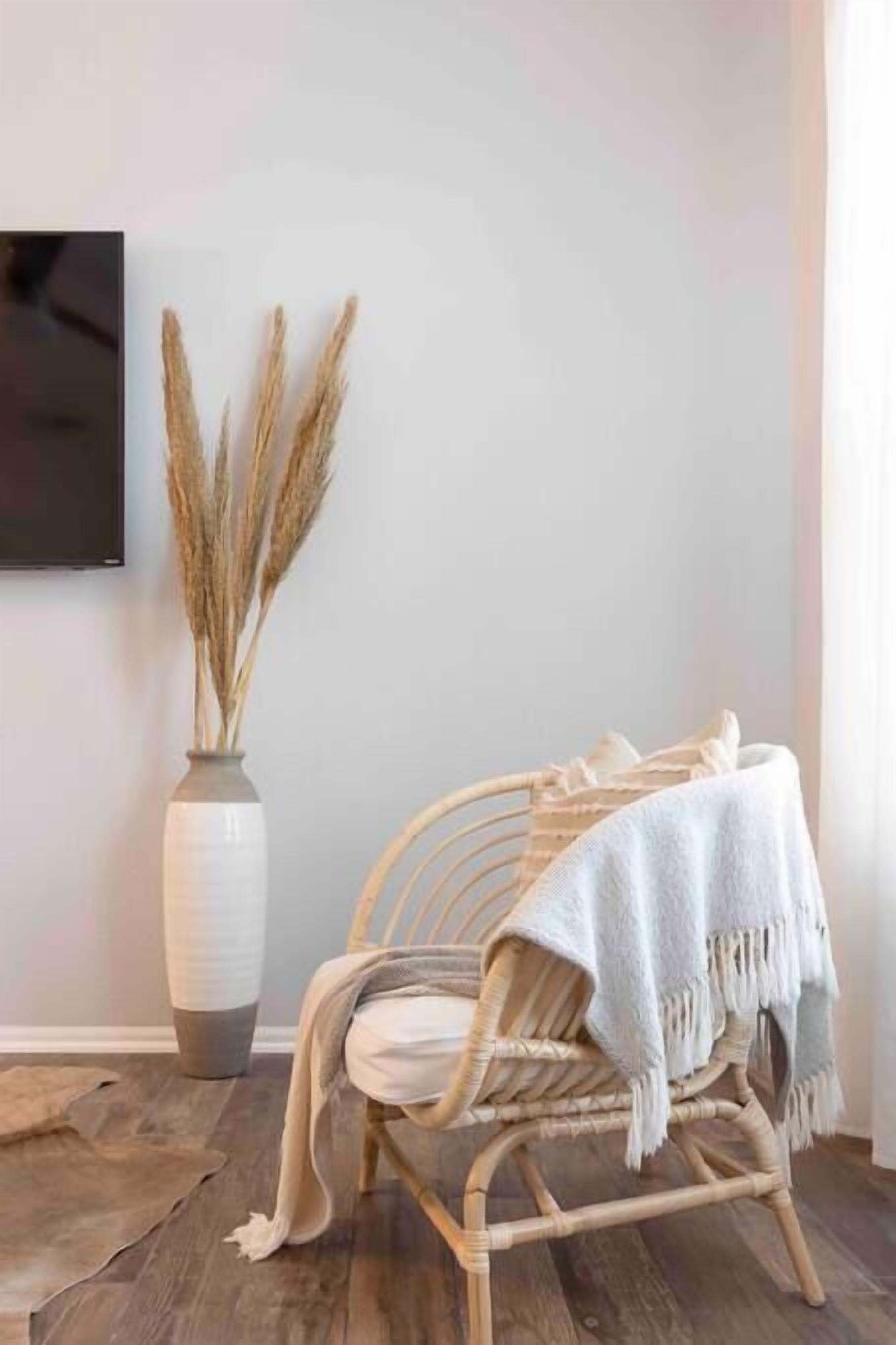Une pièce avec un fauteuil en rotin, un plaid blanc et un vase contenant des herbes des pampas séchées.