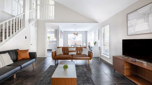 A modern living room with a staircase, a brown leather sofa, a white coffee table, and a TV on a wooden stand. - Private & Clean Retreat Near Lackland AFB | Pet-Friendly & Family Ready! (San Antonio)