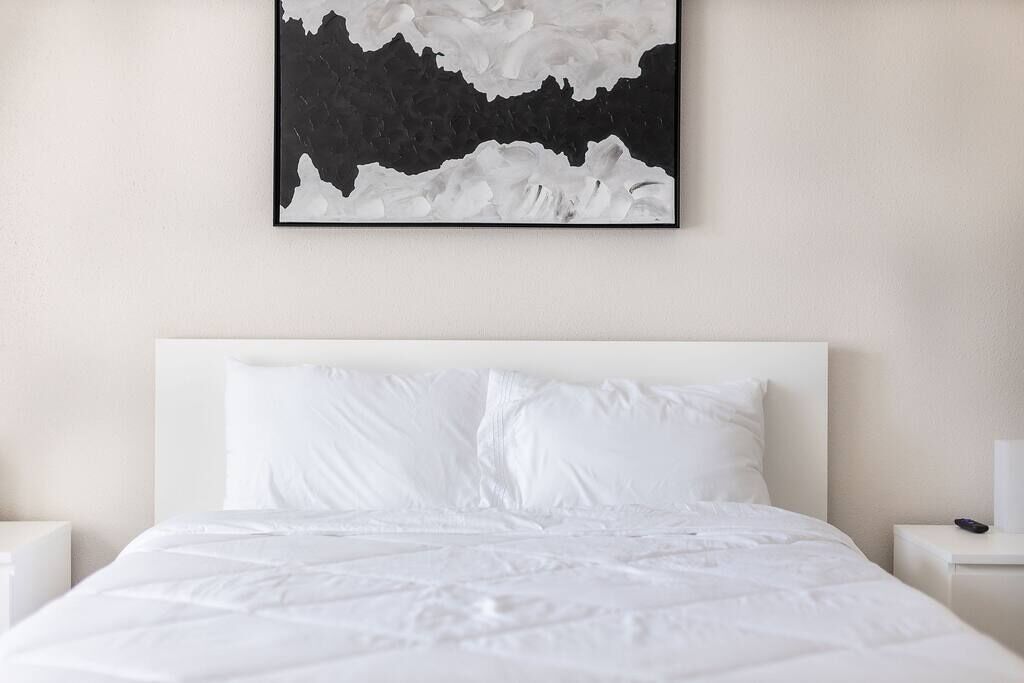 A modern hotel room with a large bed, white bedding, and a black and white abstract painting above the bed.