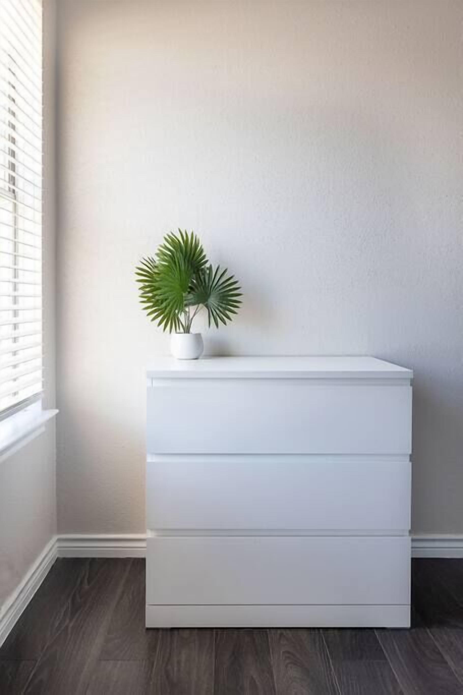 A white chest of drawers against a plain wall with a potted plant on top.