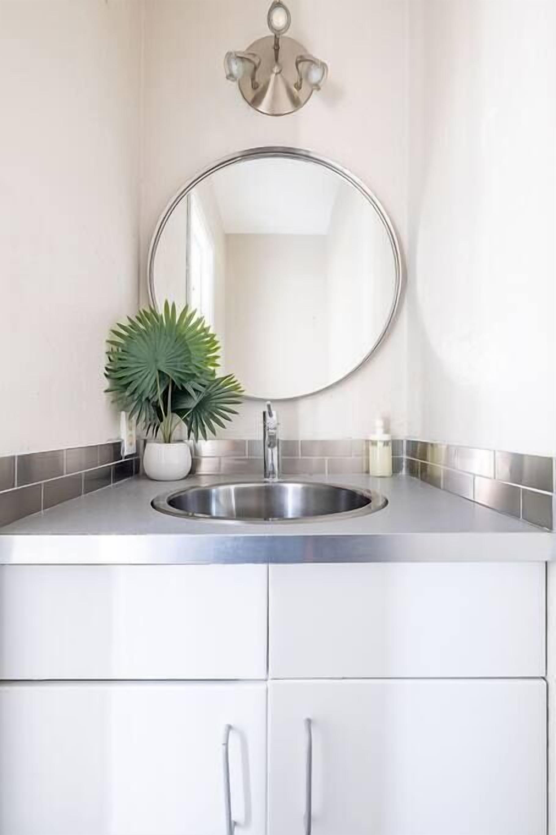 A bathroom with a round mirror, stainless steel sink, white cabinets, a potted plant, and a wall-mounted light fixture.