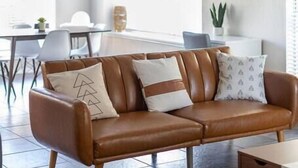 A modern living room with a brown leather sofa, a dining area with a white table and chairs, and a kitchen in the background. - Private & Clean Retreat Near Lackland AFB | Pet-Friendly & Family Ready! (San Antonio)