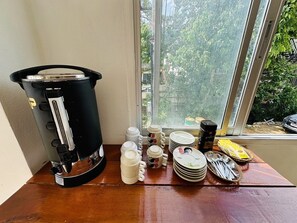 A coffee maker on a wooden table with a window view, white mugs, plates, and utensils. - Kata Place Hotel (Karon)