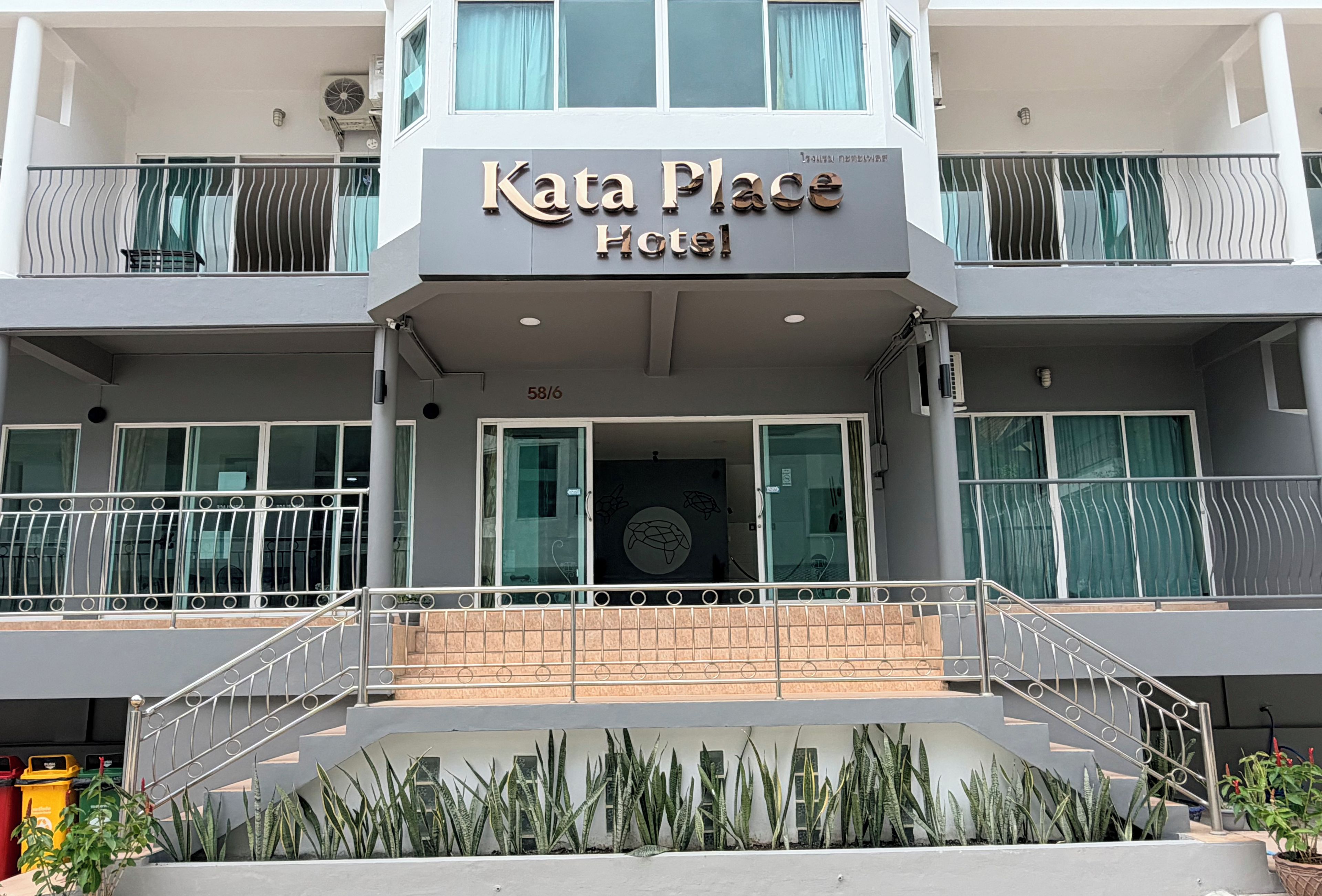 Ett hotell som heter ”Kata Place Hotel” med en modern fasad, en trappa som leder till entrén och en balkong med metallräcken.