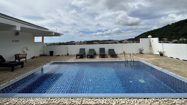 A rooftop pool with lounge chairs, a no-jumping sign, and a view of a cityscape and hills. - Kata Place Hotel (Karon)