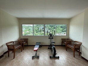 A room with a bench, an exercise bike, and a view of a green landscape through large windows. - Kata Place Hotel (Karon)