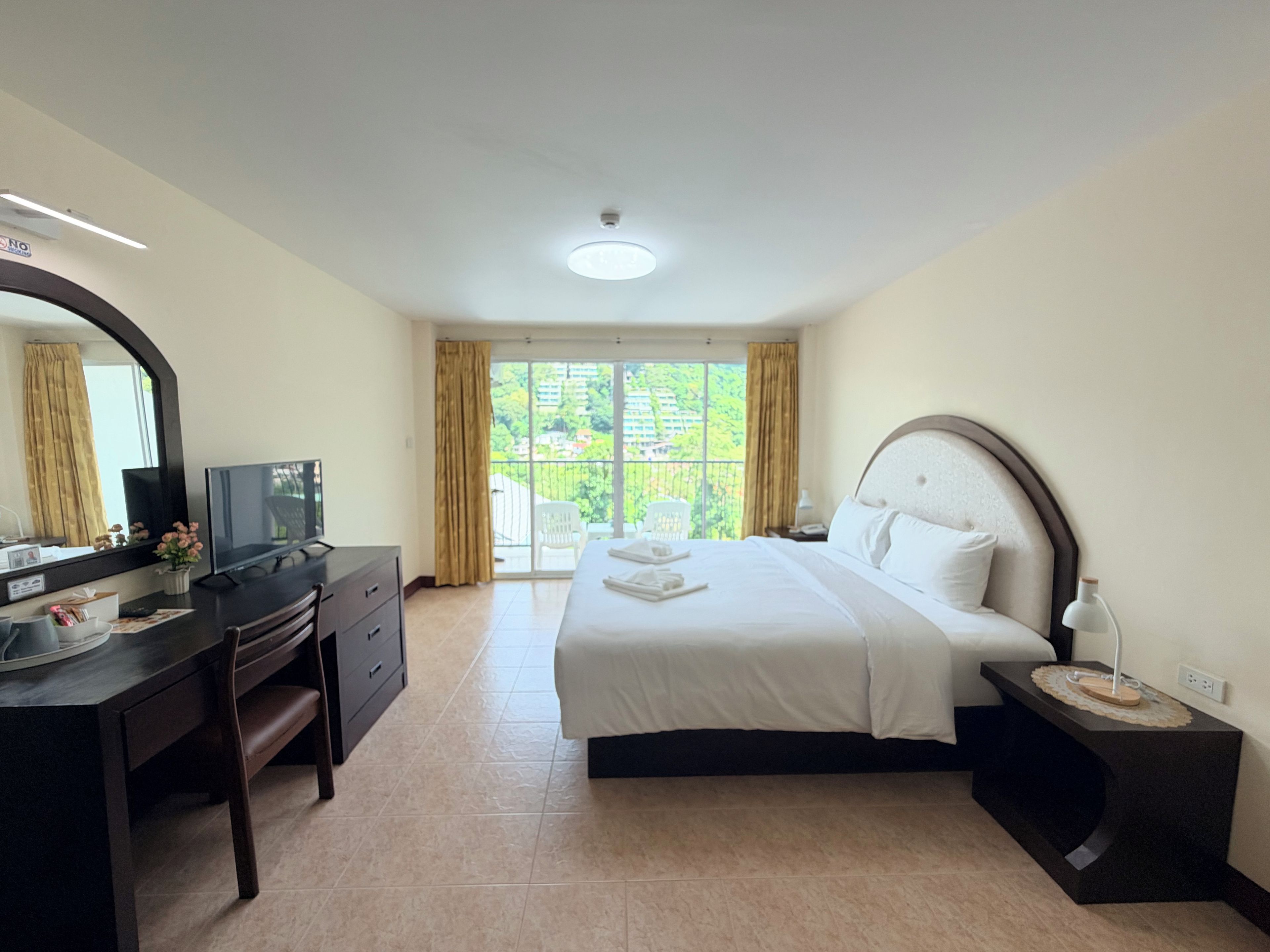 A hotel room with a bed, a desk with a TV, a mirror, and a view of greenery outside.