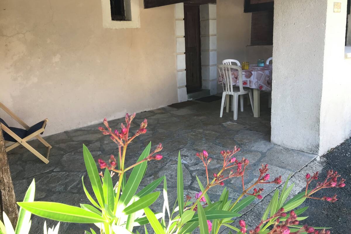 Une terrasse en pierre avec une table et des chaises, une petite fenêtre et une plante aux fleurs roses.