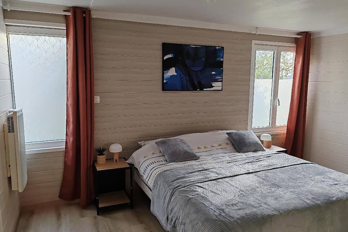 Une chambre à coucher avec un lit, une table de chevet, un téléviseur fixé au mur et une fenêtre avec des rideaux.
