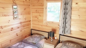 A wooden cabin room with two beds, a nightstand, a lamp, and a window with curtains. - Les Gîtes de Petrocoriis (Boulazac-Isle-Manoire)