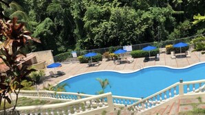 A swimming pool with a curved edge, surrounded by a white balustrade. There are lounge chairs and umbrellas under a shaded area.