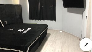 A bedroom with a bed, black curtains, a flat-screen TV mounted on the wall, and a light-colored wooden floor.