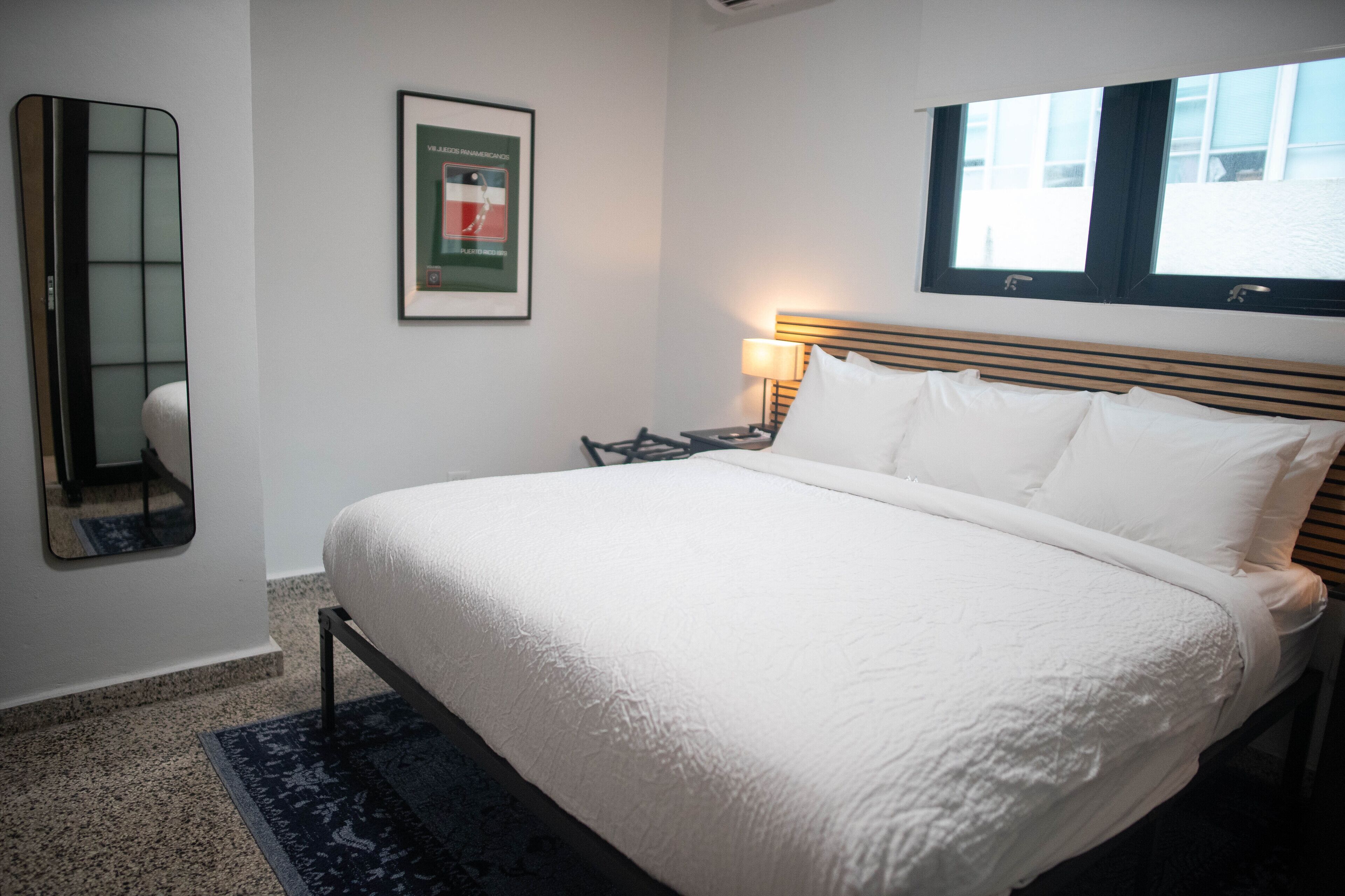 A neatly made bed with white linens, a bedside table with a lamp, a framed picture on the wall, and a window with blinds.