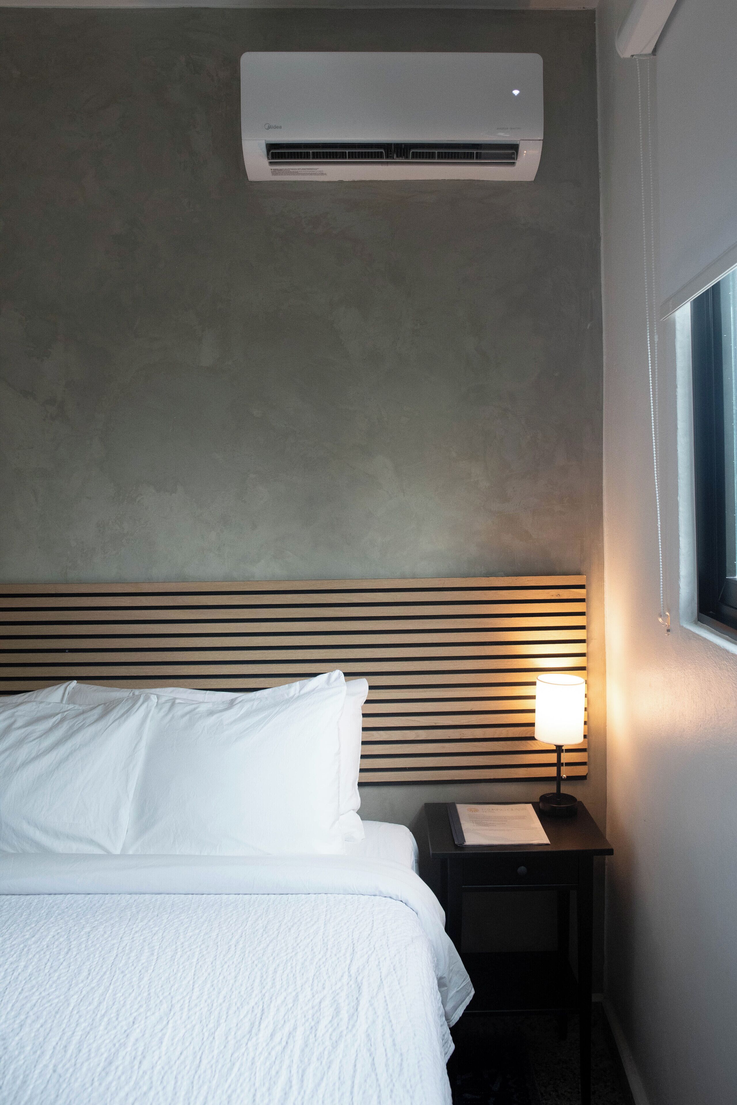 A hotel room with a bed, a nightstand with a lamp, and an air conditioning unit on the wall.