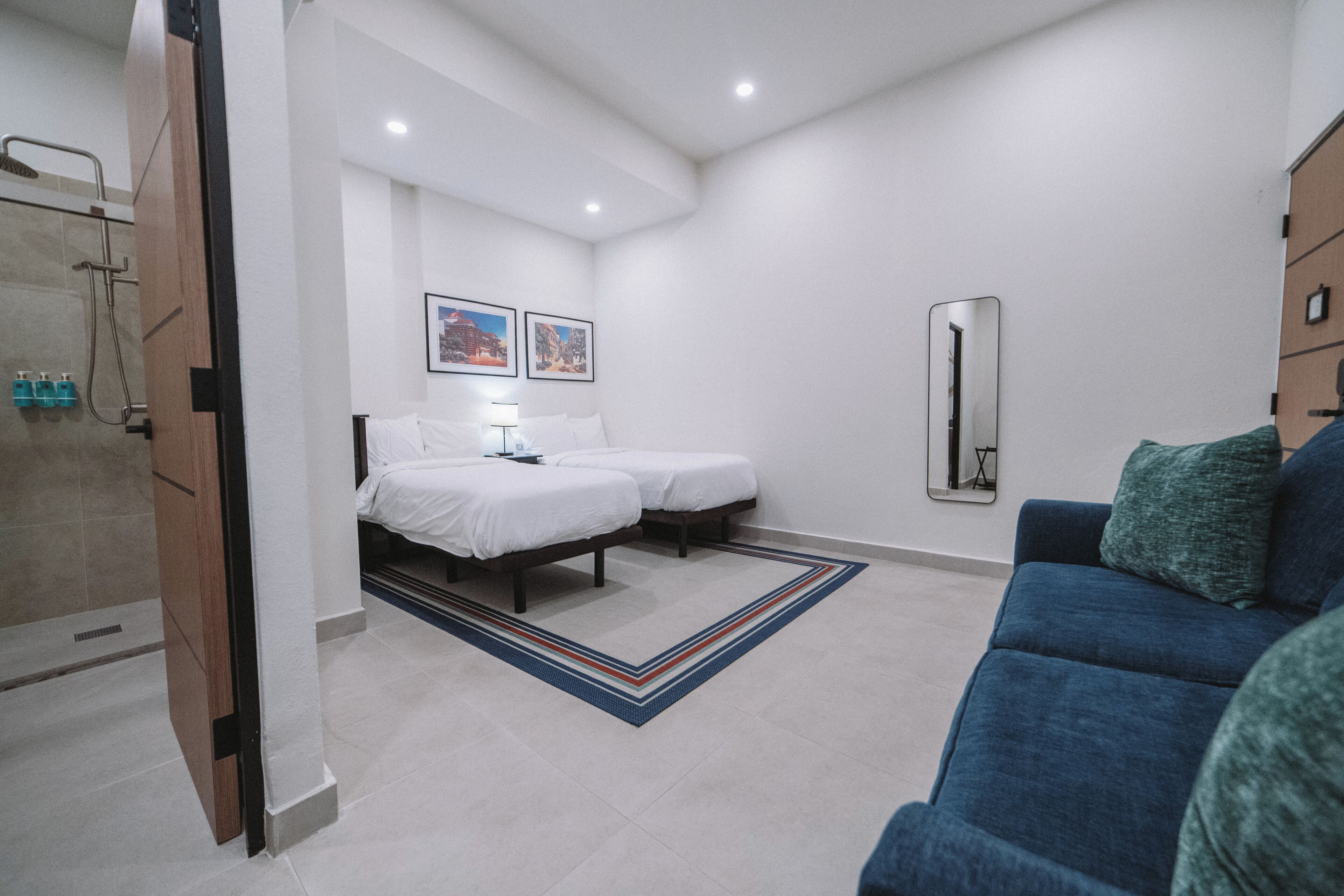 A modern hotel room with two single beds, a shower area, a blue sofa, and a mirror on the wall.
