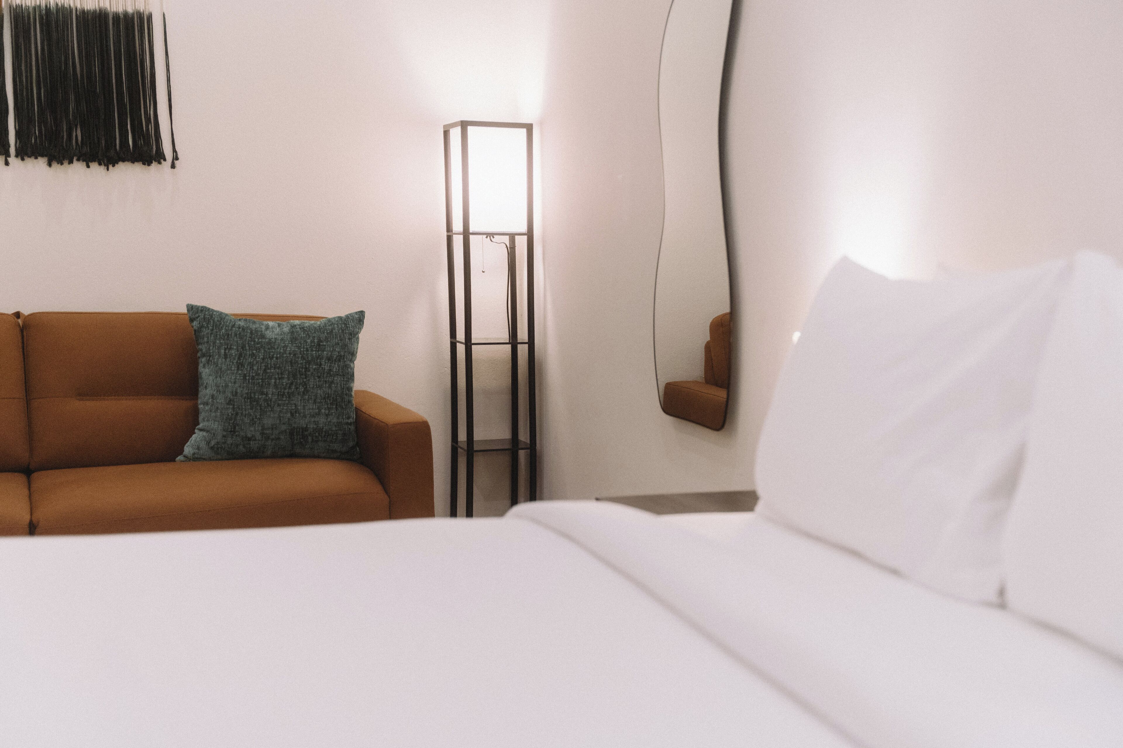 A hotel room with a brown sofa, a white bed, a floor lamp, and a mirror.