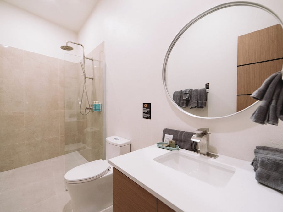 Un baño moderno con ducha de vidrio, inodoro blanco, lavamanos con espejo y toallas colgadas en un perchero.