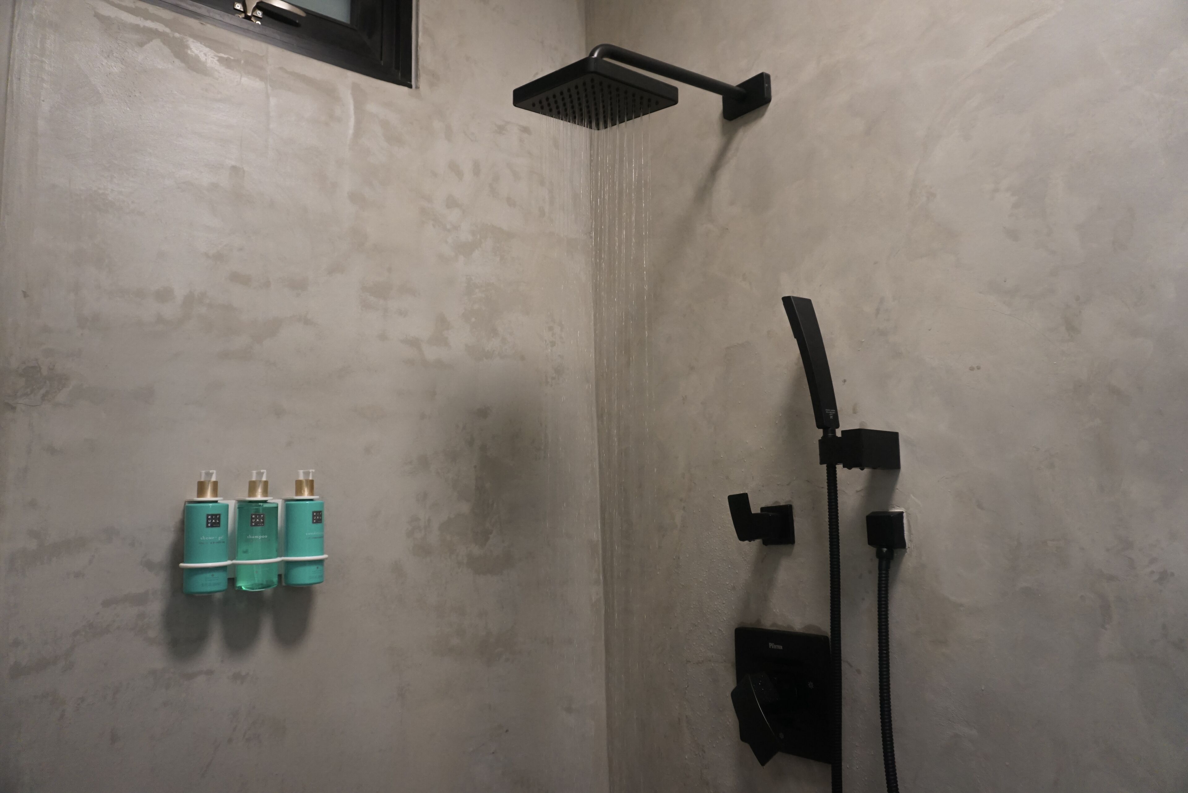 A shower area with a black handheld showerhead and a wall-mounted square showerhead. There are three bottles of shampoo and conditioner on the wall.