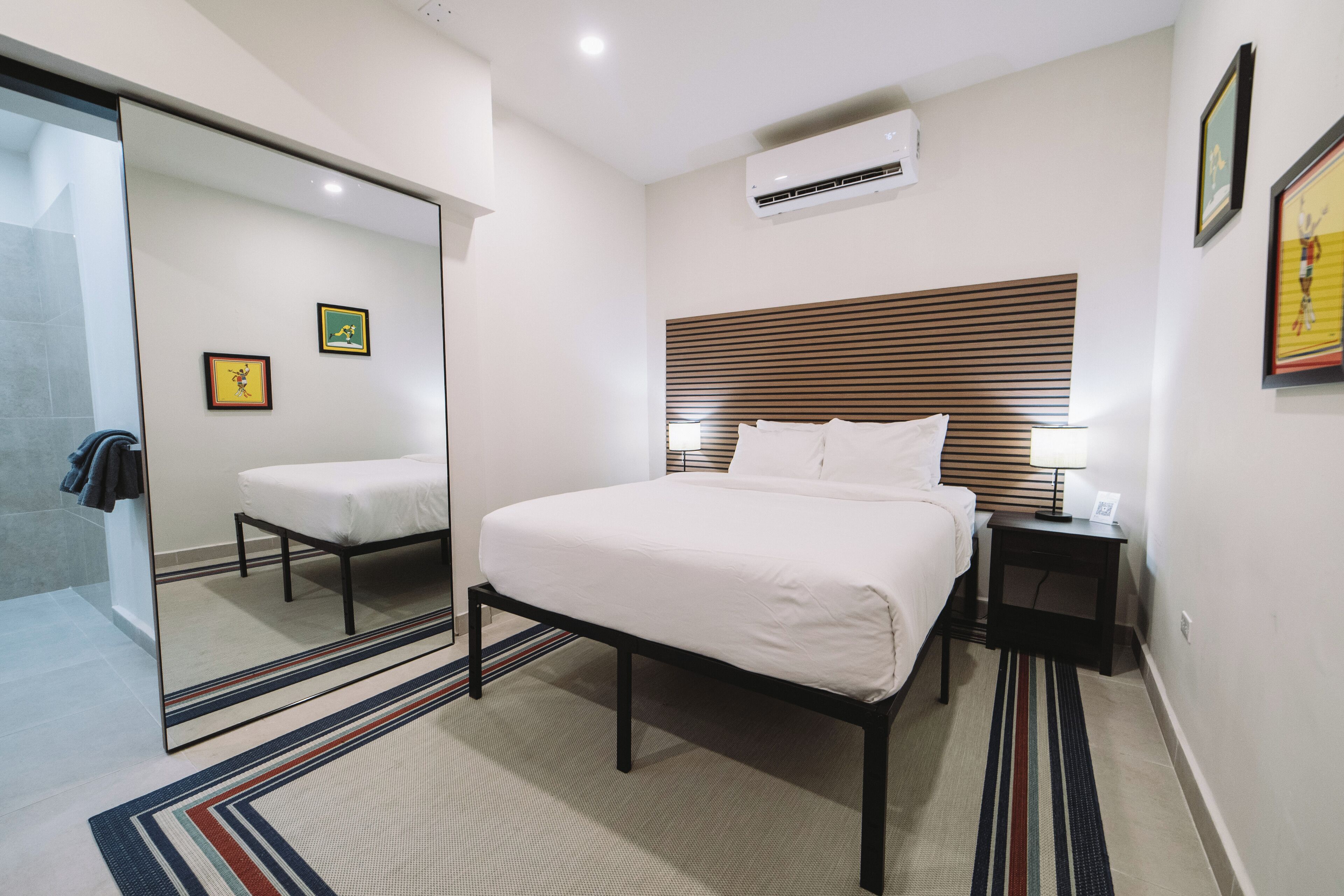 A modern hotel room with a large bed, bedside tables, a mirror, and a wall-mounted air conditioner.