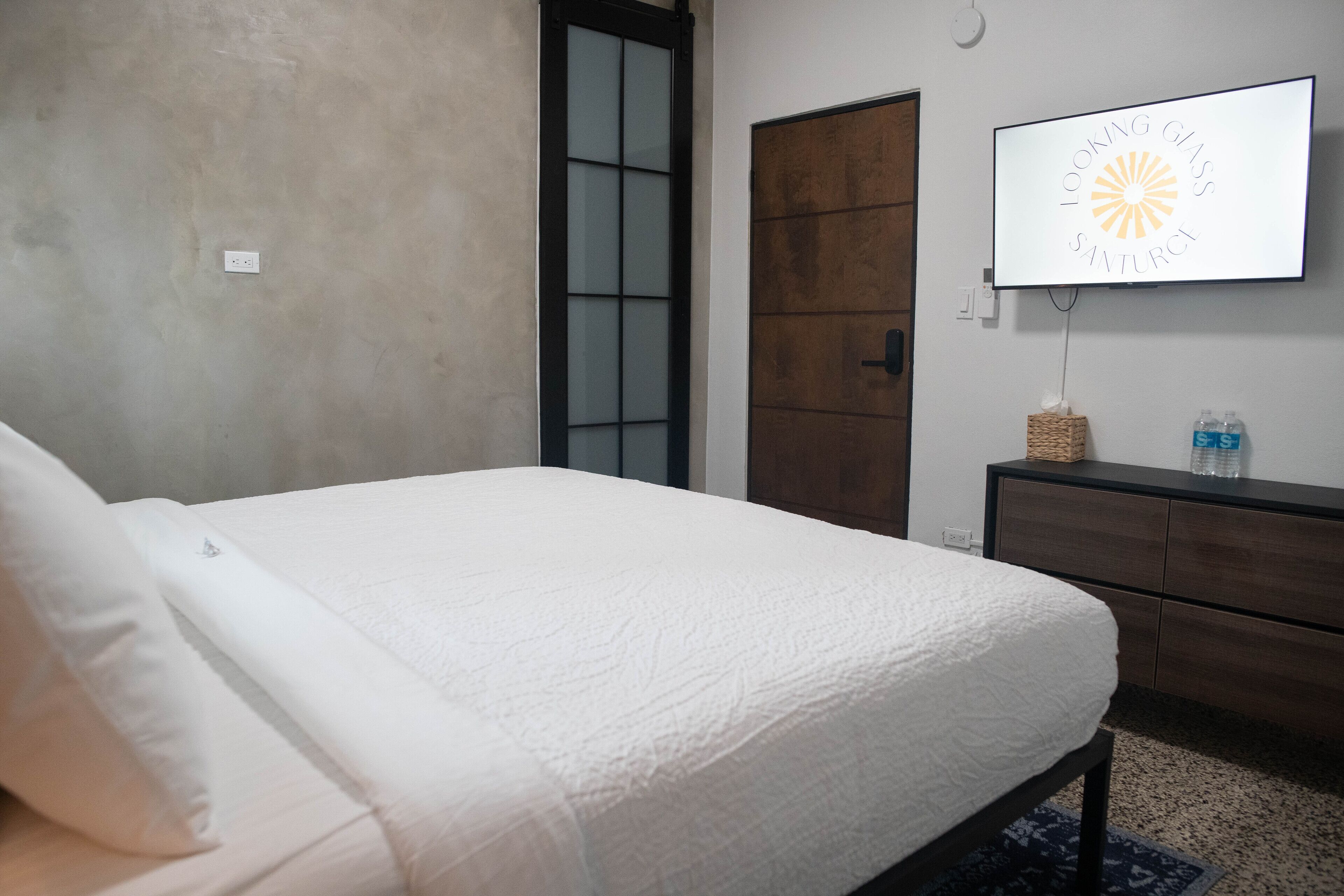 A bedroom with a bed, a wooden door, a TV on the wall, and a small dresser.
