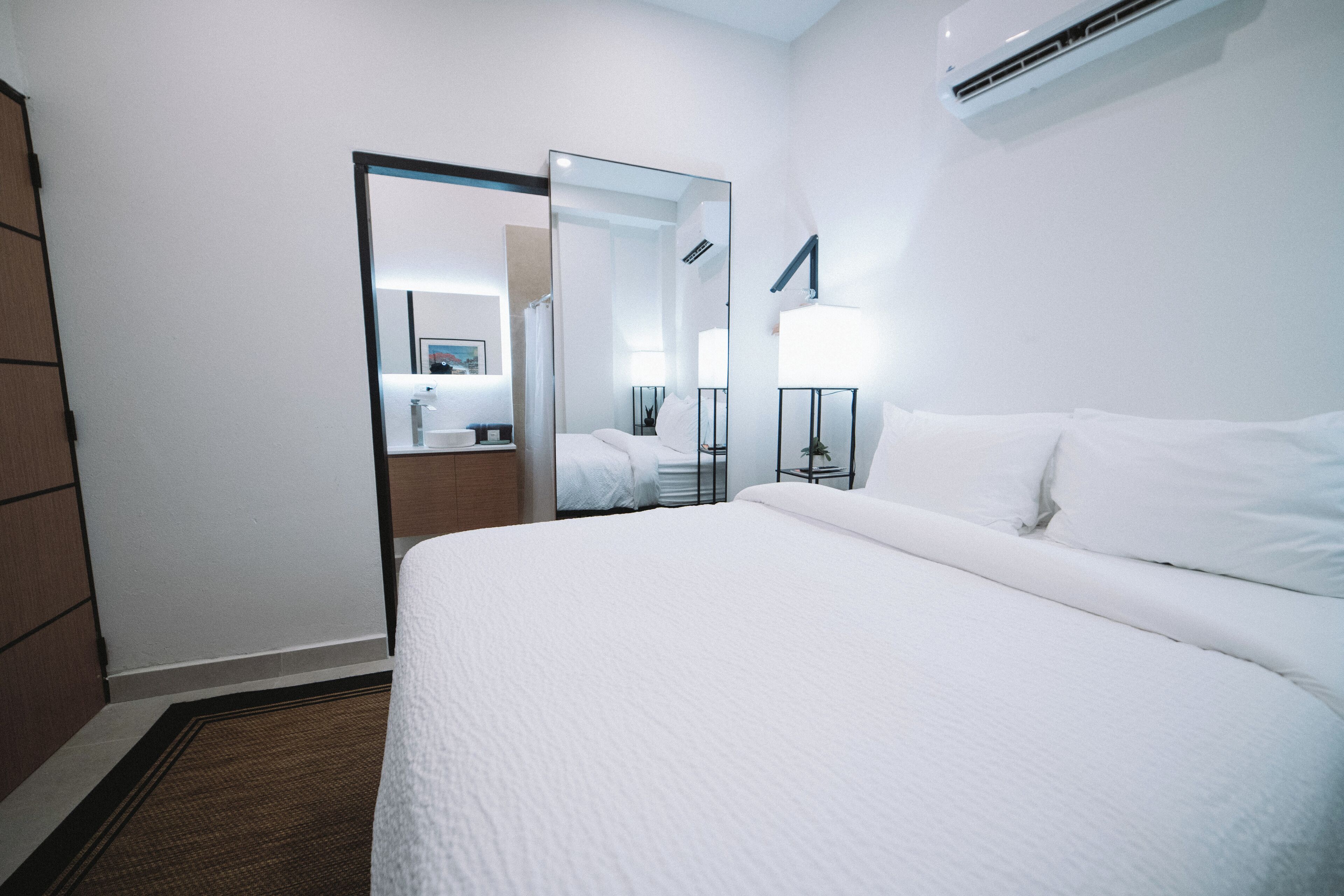 A hotel room with a large bed, a mirror, a bedside table with a lamp, and an air conditioning unit on the wall.