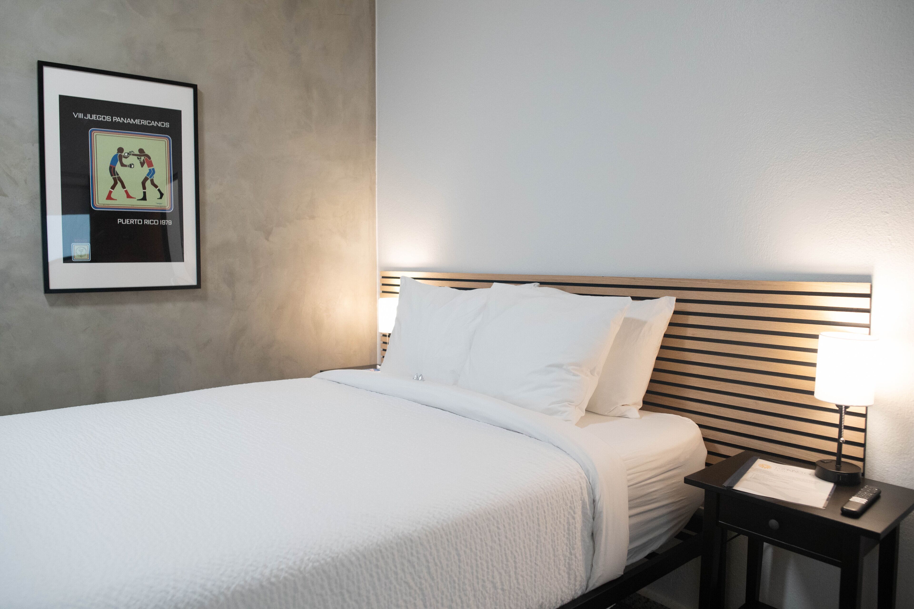 A neatly made bed with white linens, a framed picture on the wall, and a bedside table with a lamp and remote control.