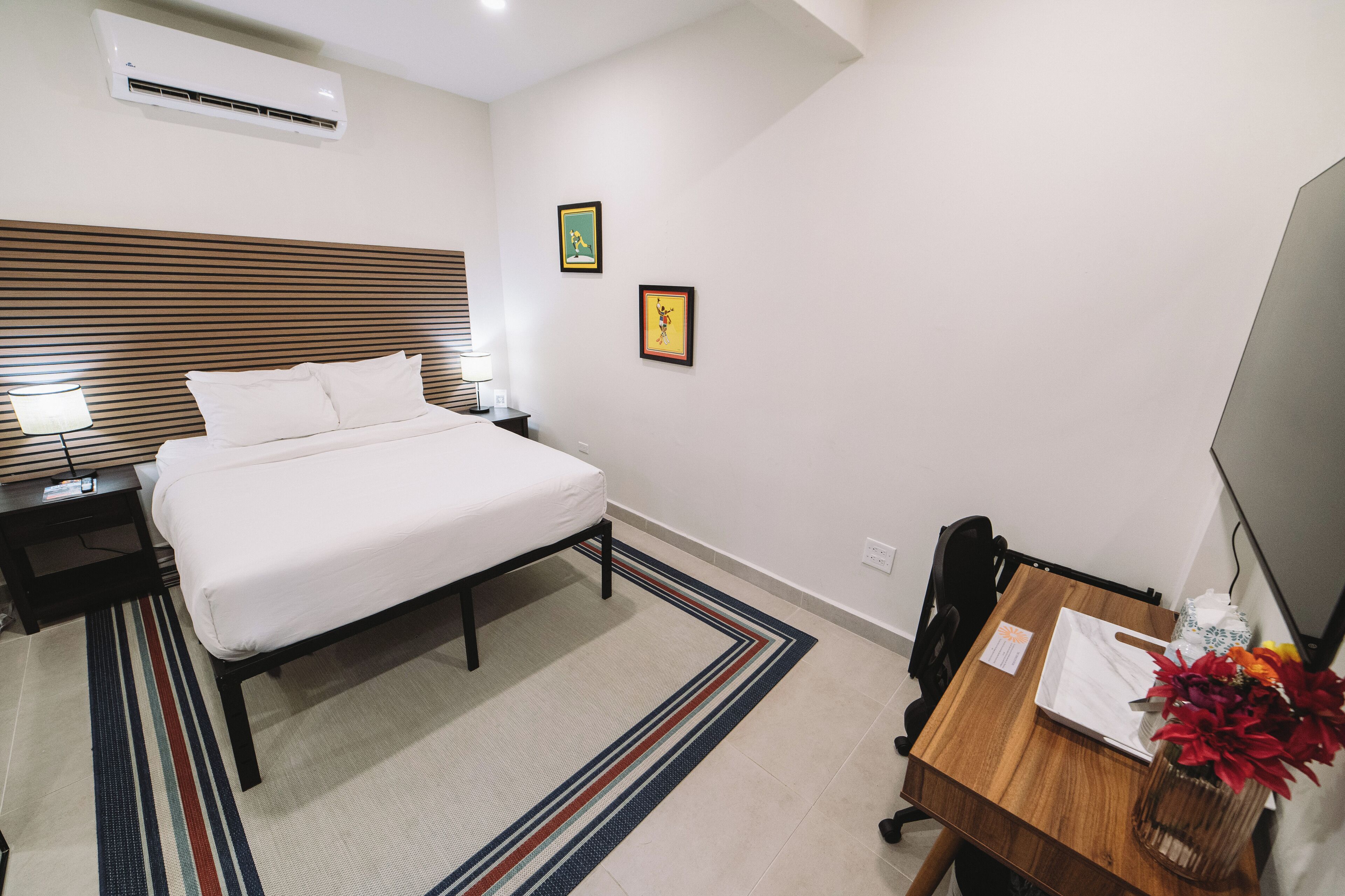 A hotel room with a bed, bedside table, lamp, and a small desk with a chair. There are two framed pictures on the wall and a vase with flowers on the desk.