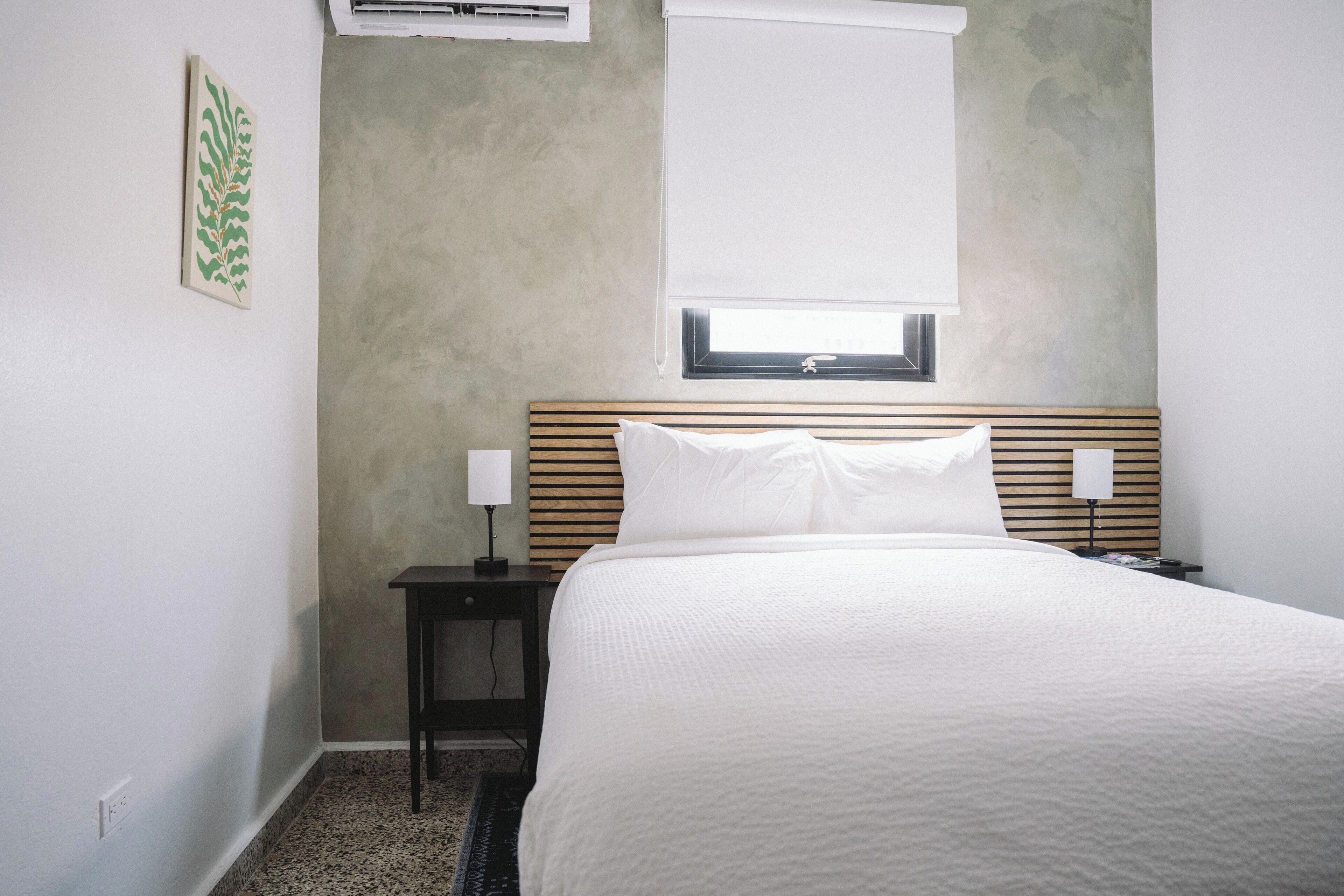 A hotel room with a bed, a nightstand, a lamp, and a window with blinds.