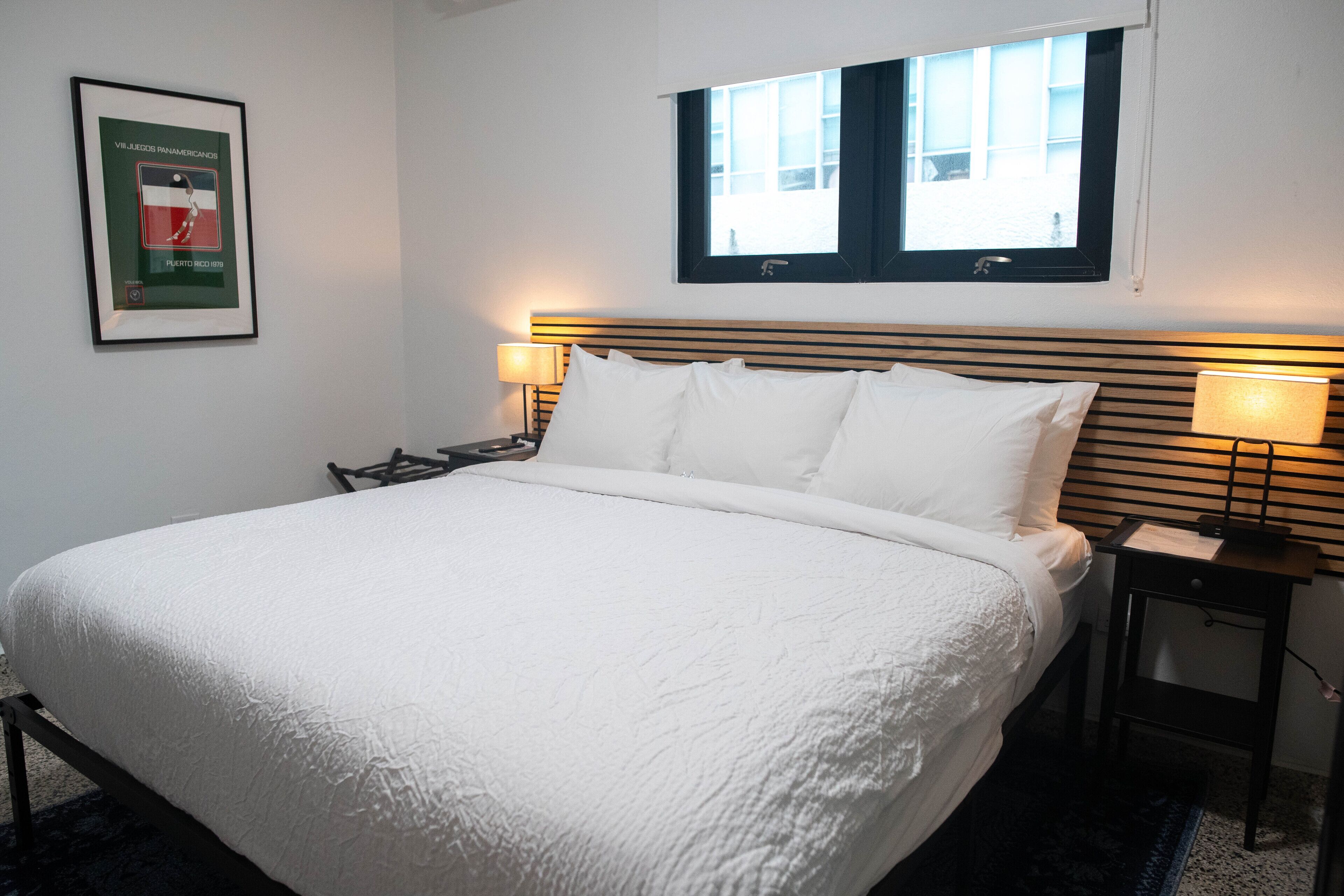 Een keurig opgemaakt bed met witte lakens in een hotelkamer. Er hangt een lijstje aan de muur, een raam met zonwering en een nachtkastje met een lamp.
