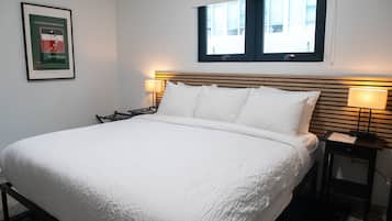 Een keurig opgemaakt bed met witte lakens in een hotelkamer. Er hangt een lijstje aan de muur, een raam met zonwering en een nachtkastje met een lamp.