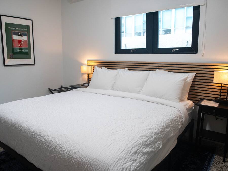 Een keurig opgemaakt bed met witte lakens in een hotelkamer. Er hangt een lijstje aan de muur, een raam met zonwering en een nachtkastje met een lamp.