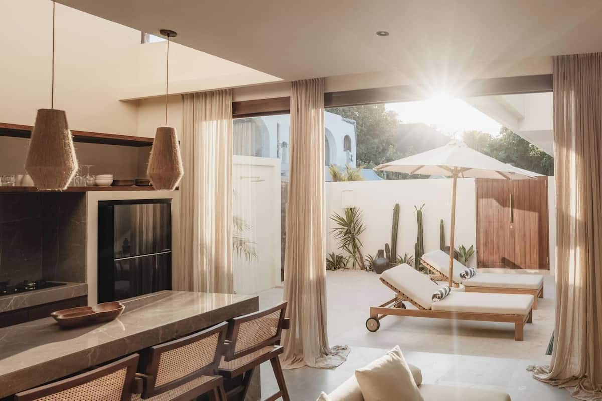 Una cocina moderna con encimera de mármol y un comedor con mesa y sillas. Hay una zona de piscina con sillones y una terraza de madera.