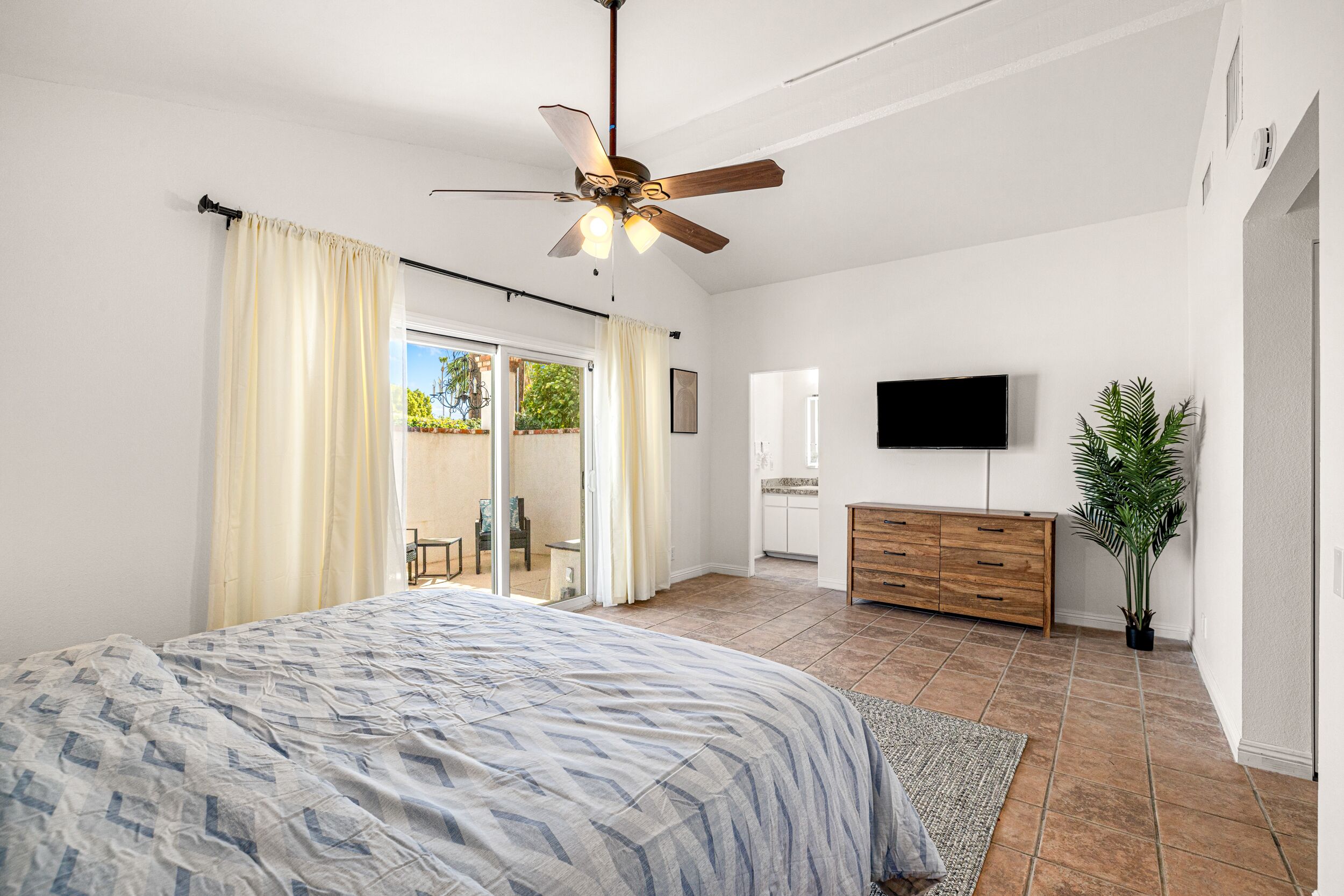 A bedroom with a bed, a ceiling fan, a TV, a plant, and a sliding glass door to a balcony.