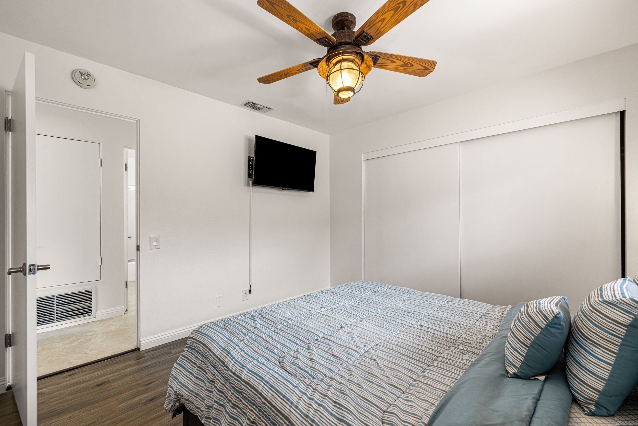 A bedroom with a bed, a ceiling fan, a mounted TV, and a door.