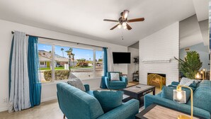 Ein modernes Wohnzimmer mit türkisfarbenen Sofas, einem Deckenventilator, einem Kamin und einem großen Fenster mit Vorhängen.
