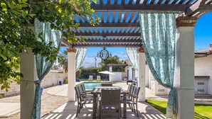 Ein überdachter Patio-Bereich mit Pool, Gartenmöbeln und einer Pergola mit gemusterten Vorhängen.